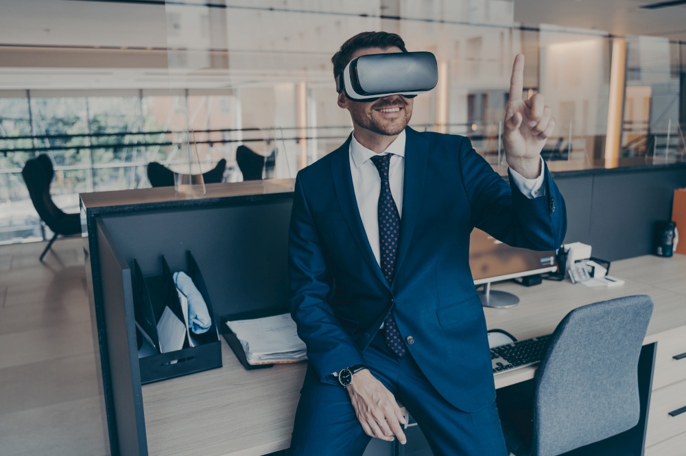 Young businessman sitting on desk in office and using VR headset on his head, pointing with forefinger in air, using gestures in app virtual interface and editing digital projects in augmented reality. Businessman sitting on desk in office and using VR headset on his head, pointing with forefinger