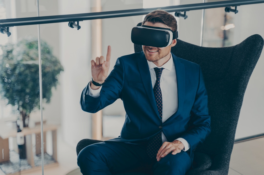 Amazed businessman in formal suit sitting in company lobby wearing virtual reality headset goggles, stunned millennial guy trying 3D experience in VR glasses, modern digital technology concept. Amazed businessman in formal suit sitting in company lobby wearing virtual reality headset goggles
