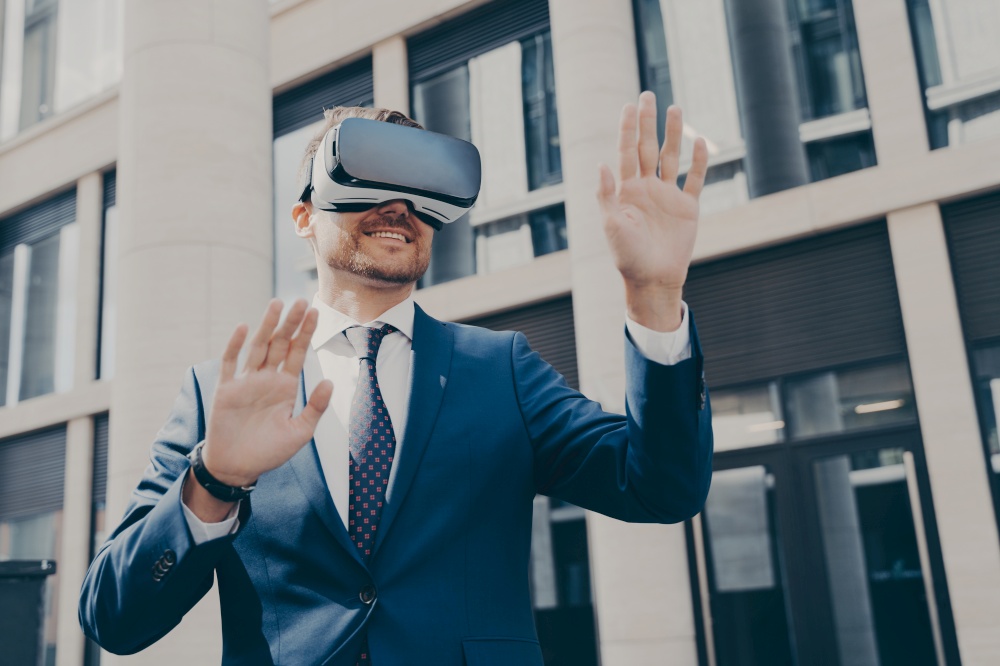 Young cheerful businessman with beard dressed in blue formal suit trying out VR glasses, checking out virtual reality, exploring digital world, standing alone next to building in background. Young businessman with beard dressed in blue formal suit trying out VR glasses outdoors