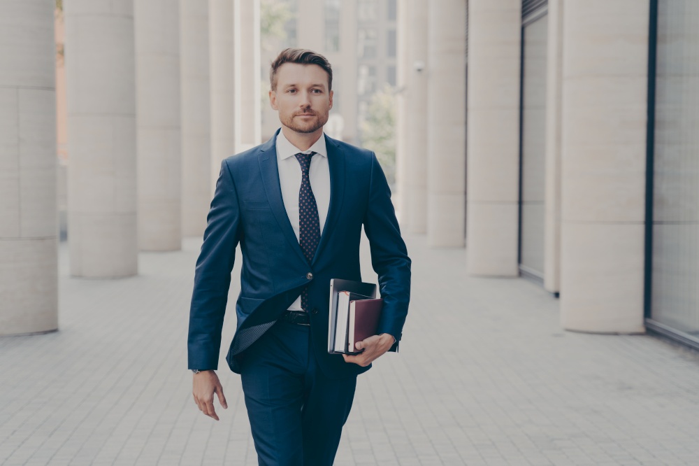 Confident successful businessman in formal clothes with laptop, notebook and book in hand walking on city street with urban background, hurrying up at business meeting with partner or client. Confident successful businessman with laptop in hand walking on city street with urban background