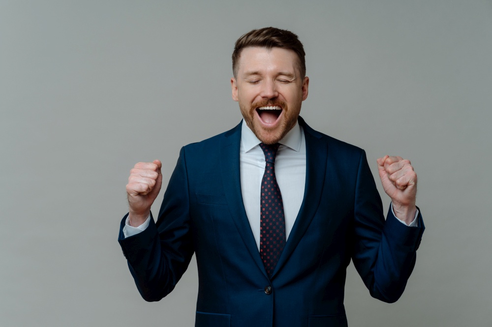 Getting good news. Excited happy businessman in elegant suit reaching business goals at work and celebrating success while standing against grey studio background, keeping eyes closed and mouth opened. Excited businessman in suit screaming and celebrating success