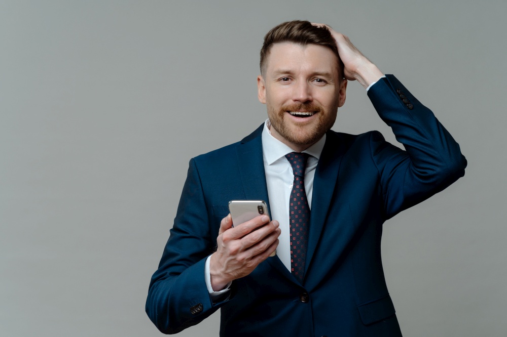 Horizontal shot of handsome bearded businessman keeps hand on head smiles gladfully holds smartphone uses platform for banking dressed formally isolated over grey background. Corporate director