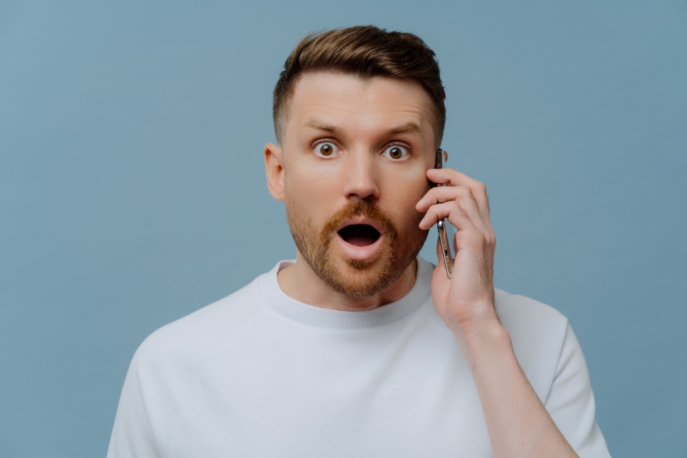 Technology and human reactions concept. Stupefied bearded handsome adult man has telephone conversation hears shocking news keeps modern smartphone near ear isolated over blue studio background.