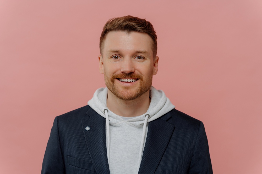 Portrait of positive attractive red haired guy wearing hoodie and jacket smiling at camera, young good looking cheerful man with beard posing in stylish casual clothes on pink studio background. Positive red haired guy on pink background