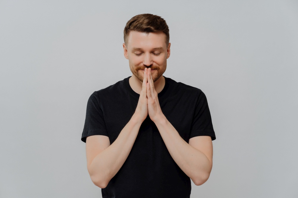Young bearded smiling man in black tshirt keeping palms pressed together and eyes closed, dreaming making wish while standing against gray background. Happy caucasian guy believing in something good. Young happy man in black t shirt with closed eyes keeping palms pressed together and dreaming about something