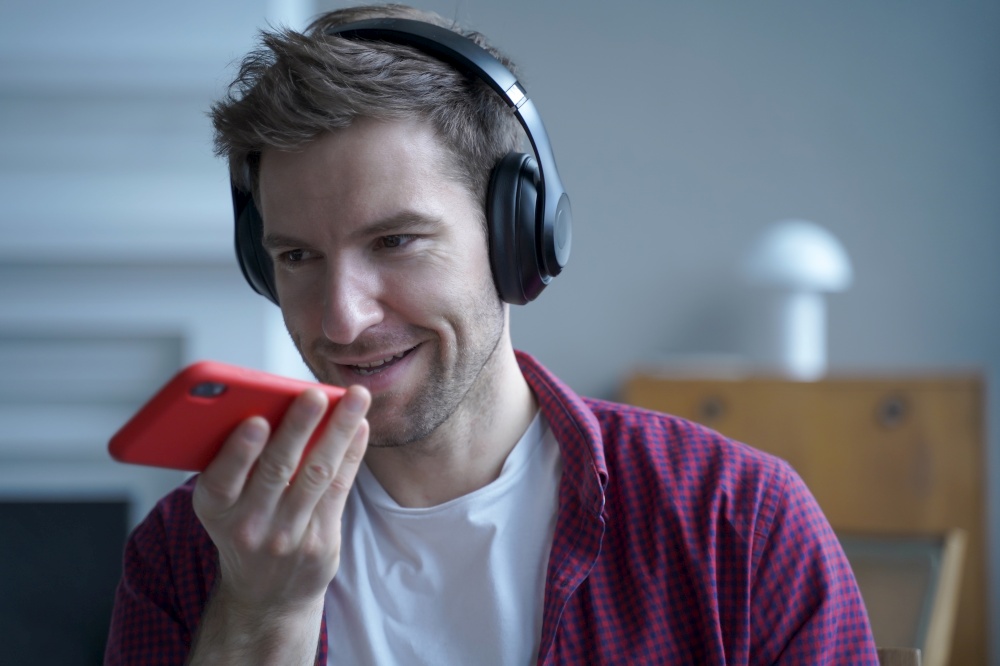 Young smiling german guy in headphones using translator app while studying online, holding mobile phone and talking on speakerphone with friend, recording voice message while spending time at home. Young smiling german guy in headphones using translator app on mobile phone while studying online