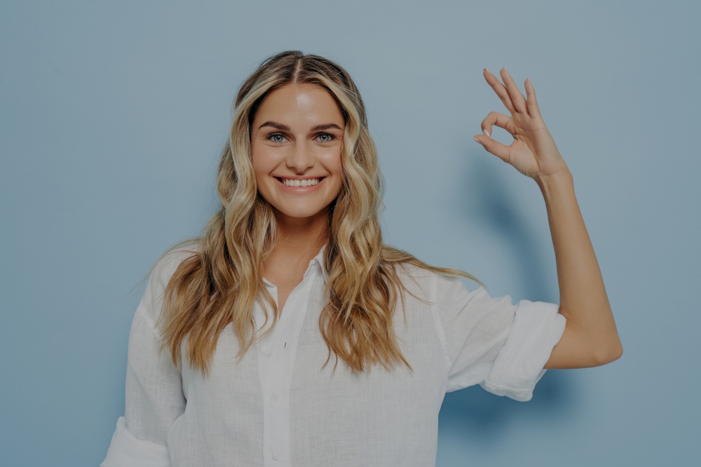 Happy smiling blonde woman showing ok gesture with her hand while smiling, looking forward to having fun and relaxing, standing next to light blue wall. Body language concept. Happy smiling blonde woman showing ok gesture