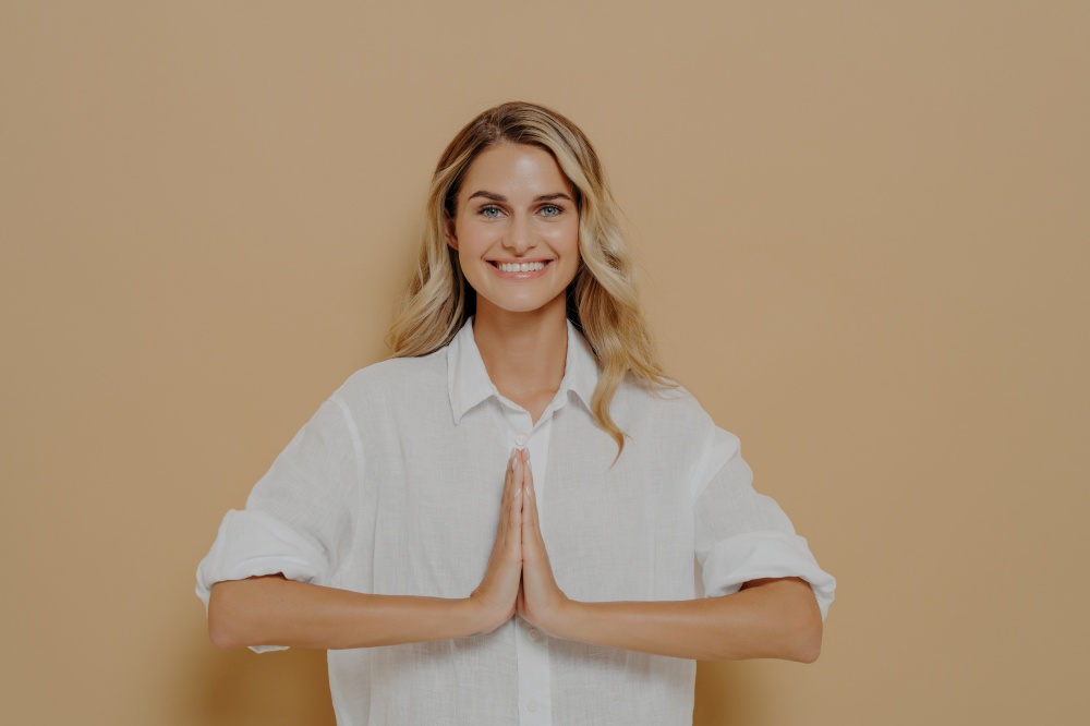Happy cute blond feamle in white shirt pressing palms together in pray, namaste or saying please gesture, smiling gratefuly and thanking for help, isolated over beige studio background. Happy cute blond girl in white shirt pressing palms together in pray or please gesture