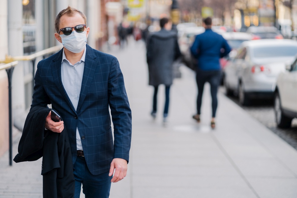 European businessman wears elegant clothes, sunglasses and protection medical mask against covid-19 or coronavirus, fights against viruses, walks through crowded city. Health and safety concept