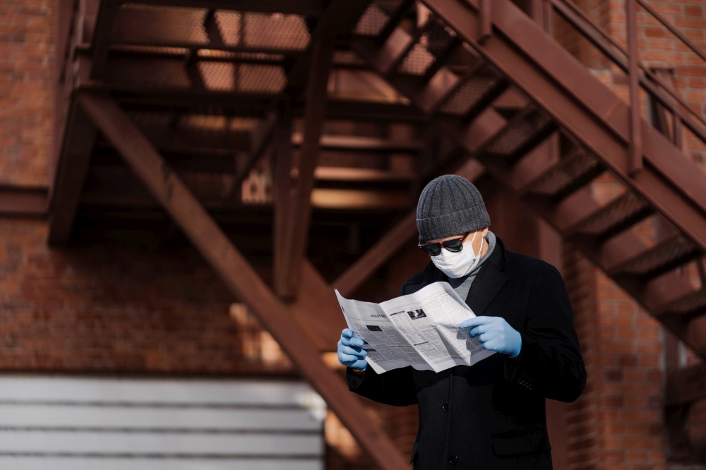 Concentrated man wears sunglasses and coat, medical mask, protective rubber gloves, reads fresh news about coronavirus spreading from newspaper, poses against blurred background with stairs.