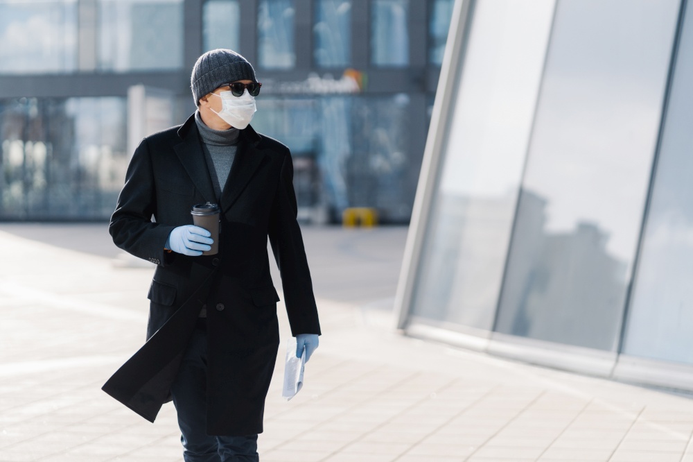 Horizontal shot of serious adult man carrying disposable cup of coffee, dressed in outerwear, protective medical mask and gloves, protects himself during coronavirus pandemic, concentrated aside