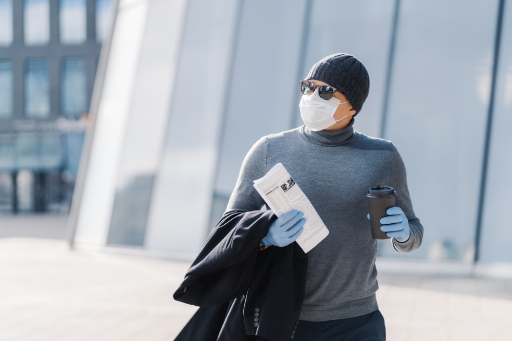Shot of man walks outdoor, protects from flu, coronavirus spreading, drinks coffee, looks aside with pensive expression, reads newspaper, follows quarantine rules. 2019-ncov. Pandemic virus.