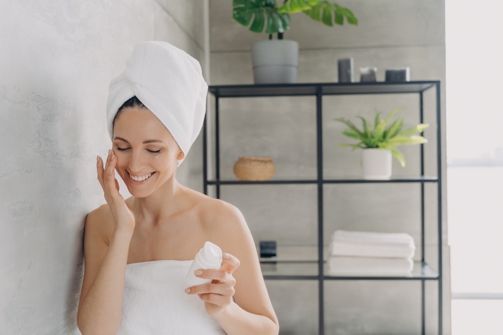 European woman applies face lotion or cream from flacon and smiling. Attractive caucasian girl wrapped in towel after bathing and hair washing. Happy young lady takes shower at home. Daily skin care.. European woman applies face lotion or cream from flacon and smiling. Daily skin care.