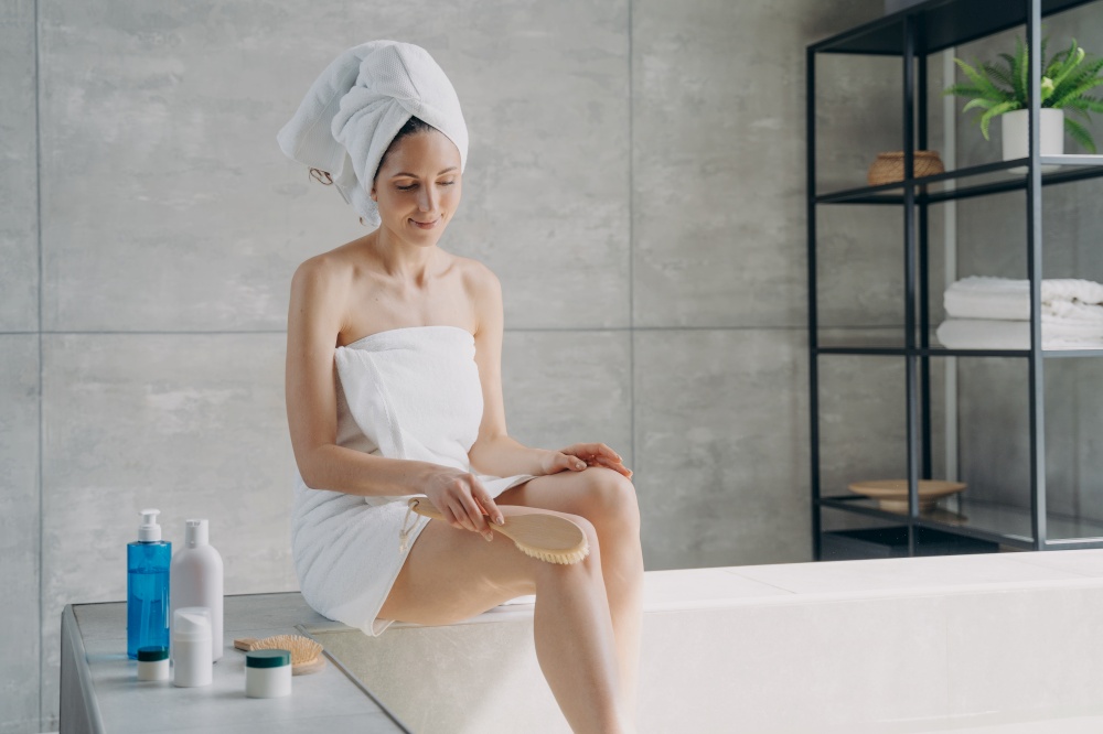 Anticellulite massage of leg with dry brush. Beautiful happy lady is making body massage with brush in bathroom. Attractive caucasian woman wrapped in towel after bathing. Girl takes shower at home.. Anticellulite massage with dry brush. Attractive caucasian woman wrapped in towel after bathing.