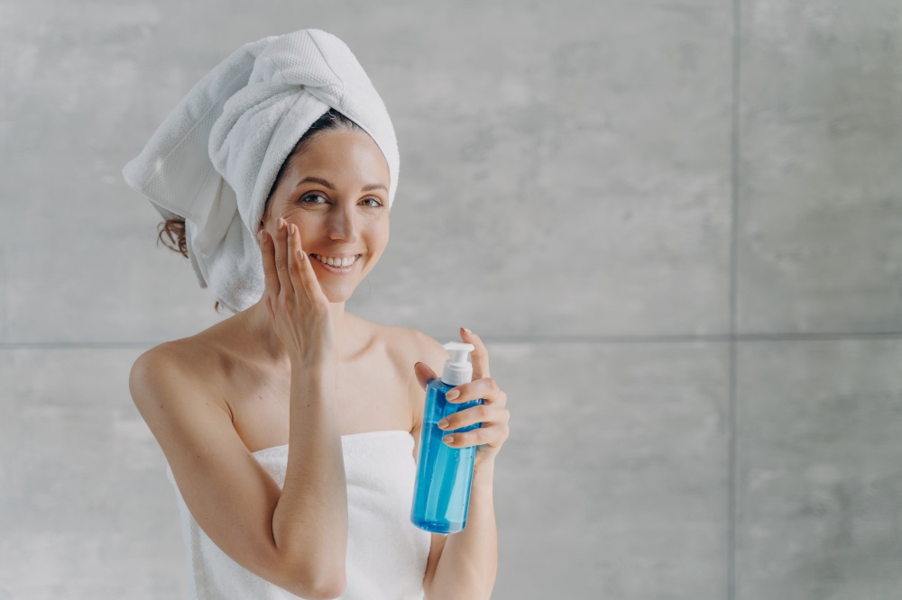 Face toner or micellar water advertising concept. Happy young woman touches pure skin. Gorgeous caucasian woman wrapped in towel after bathing applying toner in flacon. Face washing foam in bottle.. Face toner or micellar water. Gorgeous caucasian woman applying toner in flacon touching pure skin.