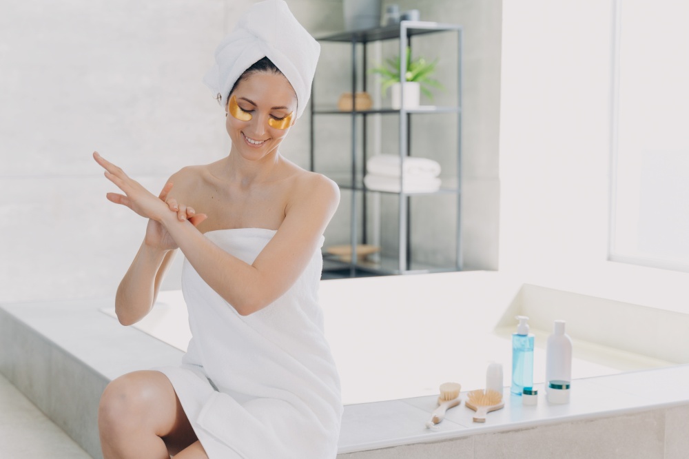 Lady applies eye patches and moisturizing skin with lotion. Attractive woman wrapped in towel after bathing is sitting on bathtub. Relaxation at spa resort. Modern interior of bathroom.. Lady applies eye patches and moisturizing skin with lotion. Relaxation at spa resort.
