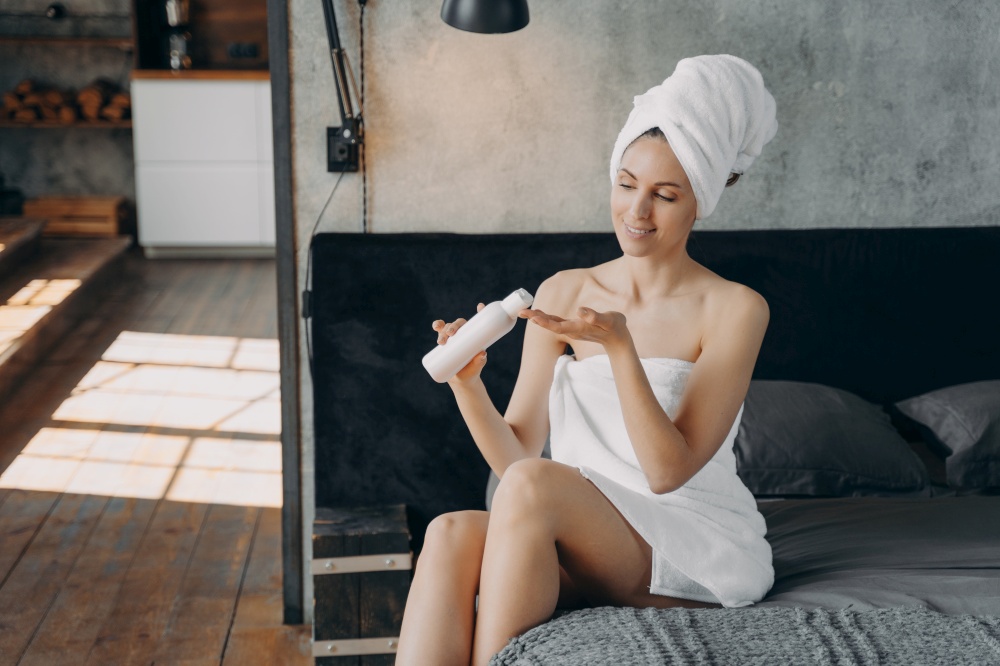 Beautiful european woman wrapped in towel is sitting in bedroom. Happy girl applying body moisturizing lotion squeezing it out from bottle after bathing. Morning shower at home.. Happy girl applying body moisturizing lotion squeezing it out from bottle after bathing at home.