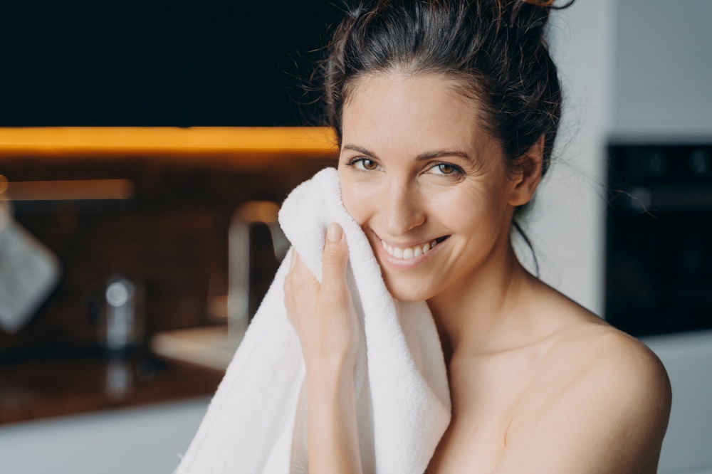 Caucasian woman is wiping face with towel after washing and smiling. Young woman takes shower at home and doing daily skin care. Hygiene and wellness, dermatology and skin cleansing.. Caucasian woman is wiping face with towel and smiling. Hygiene, dermatology and skin cleansing.
