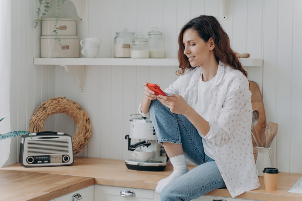 Spanish young woman typing on smartphone. Happy relaxed girl with telephone sitting on worktop of table at kitchen alone. Modern luxurious scandinavian interior. Lifestyle concept.. Spanish woman typing on smartphone. Girl sitting on worktop of table at kitchen alone.
