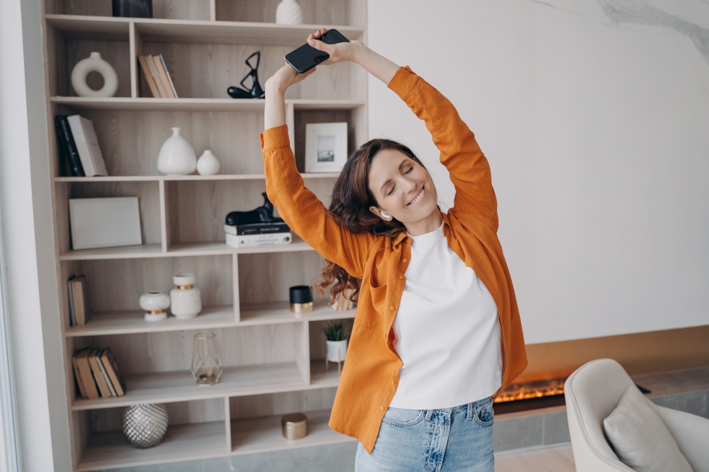Happy european woman is dancing alone at home. Girl in earphones is having fun in her living room. Music listening online. Concept of weekend, relaxation and emotion expression. Cosy home interior.. Happy european woman is dancing alone at home. Weekend, relaxation and emotion expression.