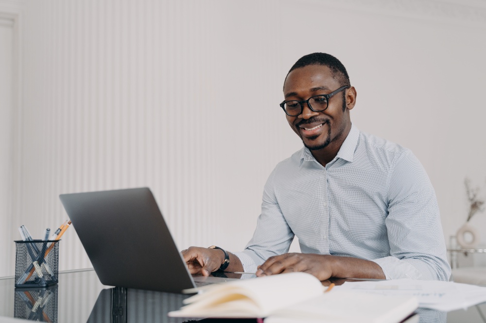 Pensive african american man wearing glasses working on laptop online, sitting at office desk, satisfied with his business project. Smiling black guy employee happy with his good job at workplace.. African american man working on laptop online satisfied with his business project, good job, smiling
