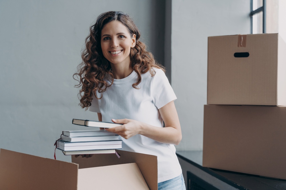 Excited girl in residential room. Happy european woman opening and unpacking boxes. Curly student in white t-shirt indoors. Relocation and moving to college concept.. Excited student in residential room. Happy european woman opening and unpacking boxes.