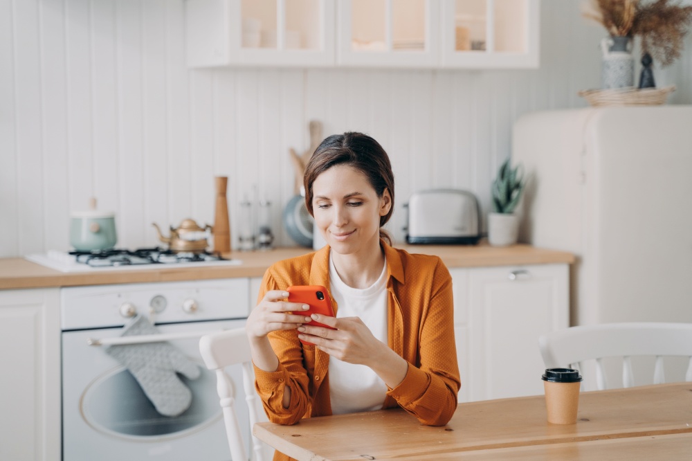 Happy european girl is sitting at the table at kitchen and texting on phone. Young woman in orange shirt is remote worker. Lady has coffee and browsing through internet. Workplace at home.. Happy girl is sitting at the table at kitchen and texting on phone. Workplace at home.