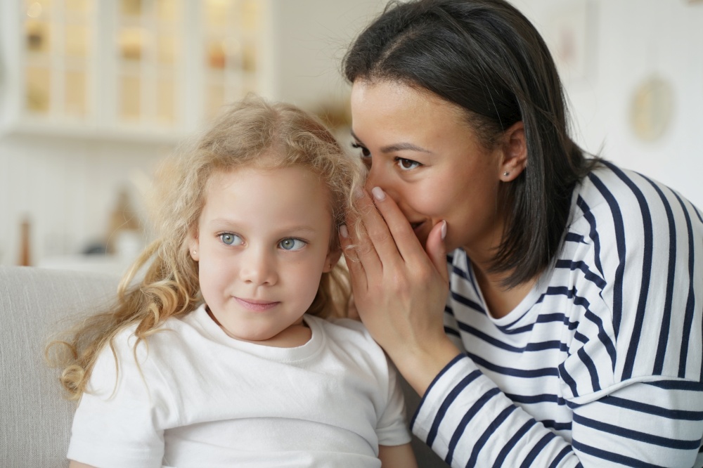 Female babysitter or mother whispering sharing secret with small kid daughter. Happy mom whisper in girl&rsquo;s ear, telling funny story to child, enjoying friendship, trust. Trusting family relationships.. Mother or babysitter whispering sharing secret with kid daughter. Trusting relationship, friendship