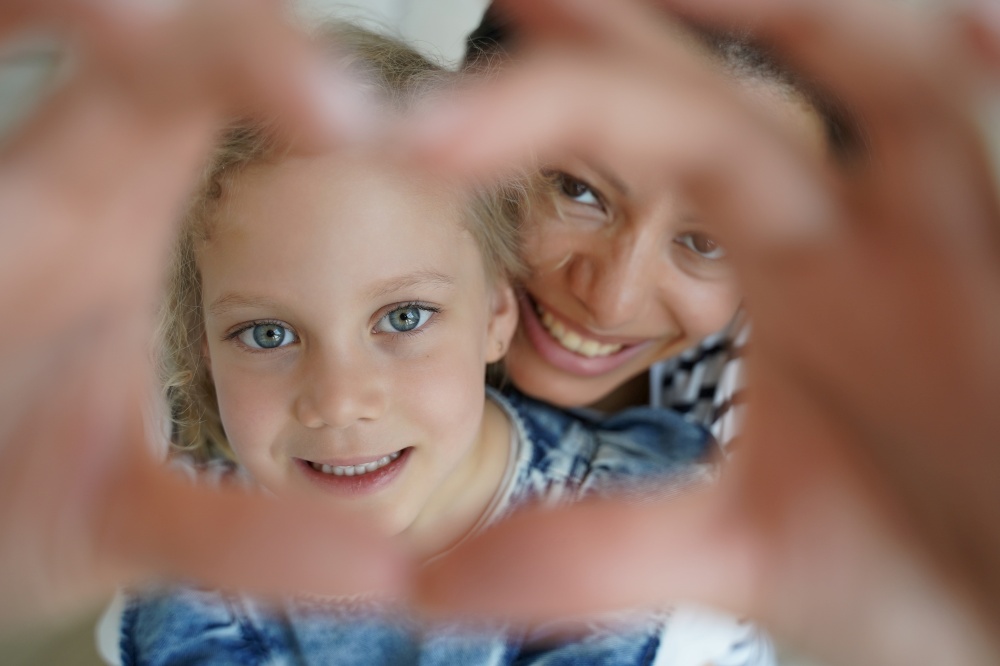 Daughter and mother make heart gesture, symbol of mom and child love care support. Smiling mum and adopted preschooler kid girl looking at camera together, show sign of gratitude. Motherhood, adoption. Adopted daughter, mother make heart gesture, symbol of child love care support. Motherhood, adoption