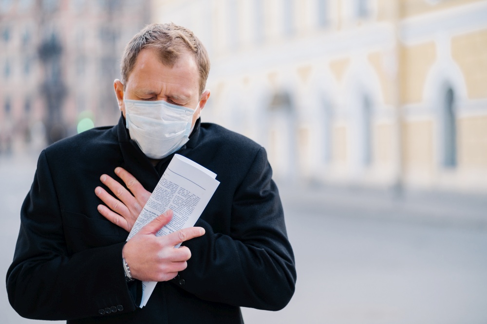 Infected man wears medical mask, has constant coughing, symptoms of Covid-19, holds rolled up newspaper, poses at city outdoor, needs isolation to stop spreading Coronavirus. Preventive measures