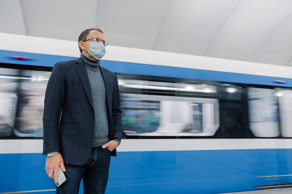 Pensive businessman wears surgical sterile mask to prevent spread of respiratory disease, poses against underground train, travels in public transport, holds smartphone in hand. Coronavirus outbreak.