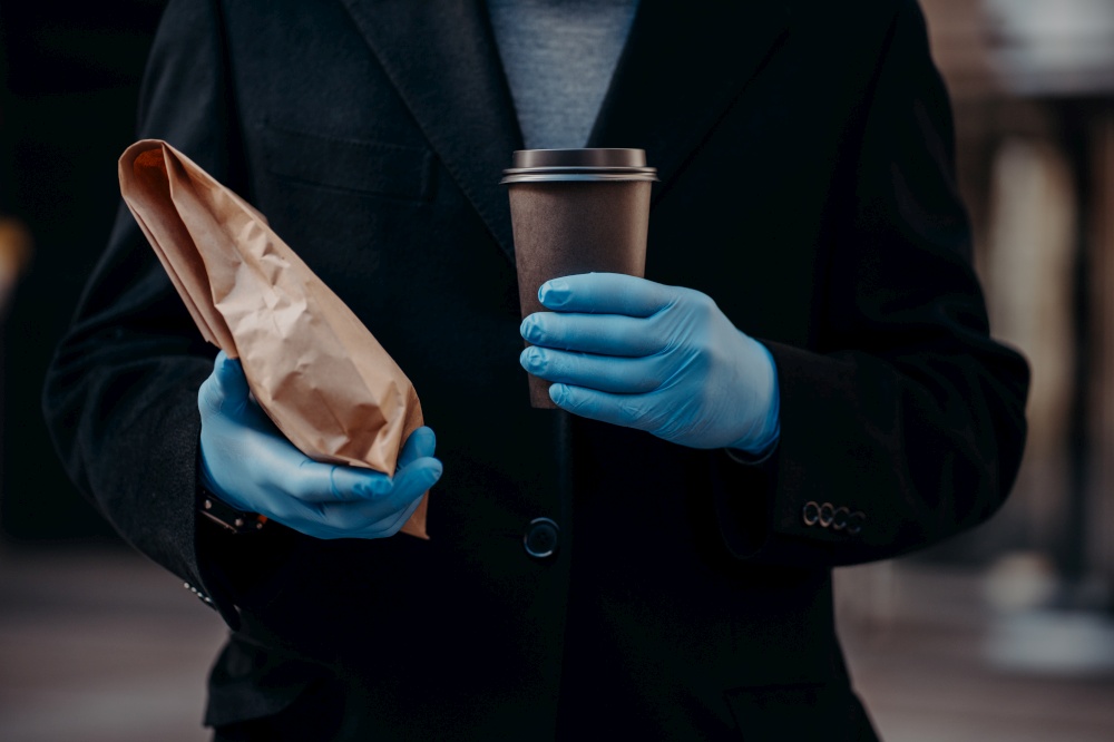Delivery service during coronavirus and quarantine. Faceless businessman holds take away food and disposable cup of coffee, wears medical rubber gloves for safety. Nutrition during pandemic.