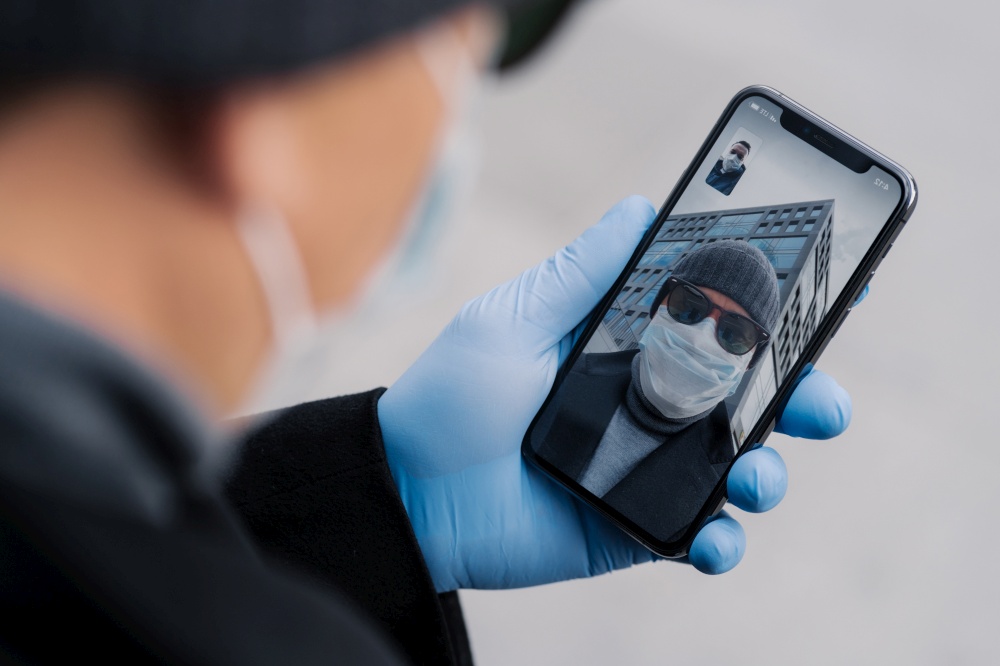 Shot of man has video conference via modern smartphone, being on distance from people during coronavirus spreading, wears protective mask and rubber gloves while walks outdoor in public place
