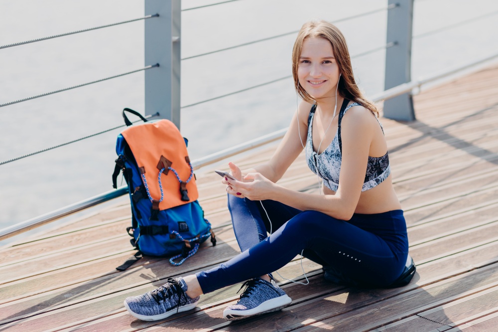 Glad sportswoman in top, leggings and sneakers has gentle smile, listens favourite music in headphones, enjoys rest after cardio training, her rucksack stands near. People and lifestyle concept