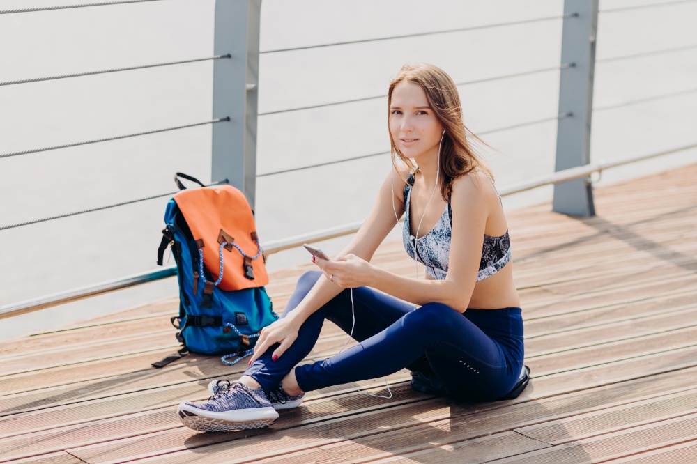 Relaxed lovely woman with dark hair, healthy pure skin, dressed in sportswear, holds cell phone for communication, listens favourite track, rucksack stands near her. People, sport, recreation concept