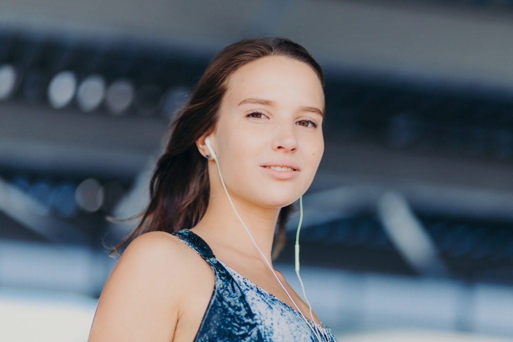 Close up shot of good looking healthy European woman listens music with unrecognizable device, looks directly at camera, poses against blurred background. People, youth, entertainmet concept