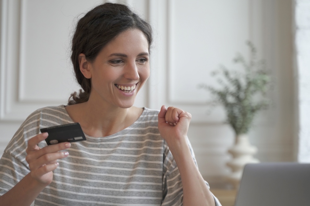 Young smiling italian female holding plastic credit card, sitting at table with laptop and shopping online or making internet payment, happy satisfied young woman using banking system on computer. Young smiling italian female holding plastic credit card, shopping online or making internet payment