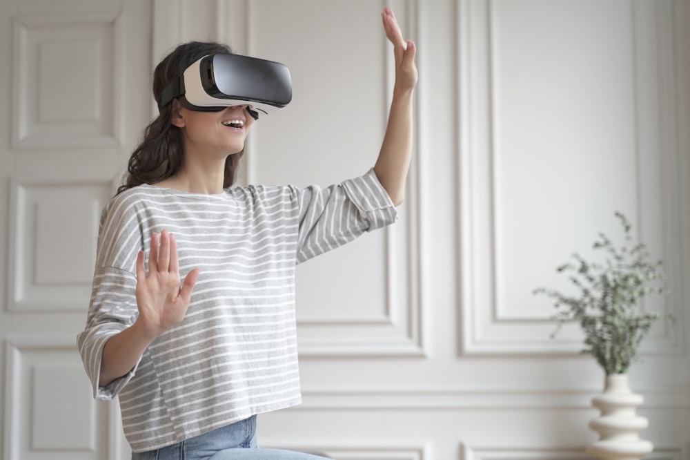 VR In eCommerce. Young happy woman in casual clothes standing indoors in virtual reality headset and enjoying fun shopping experience in augmented world, touching objects with hands. Young happy woman in virtual reality headset enjoying fun shopping experience in augmented world
