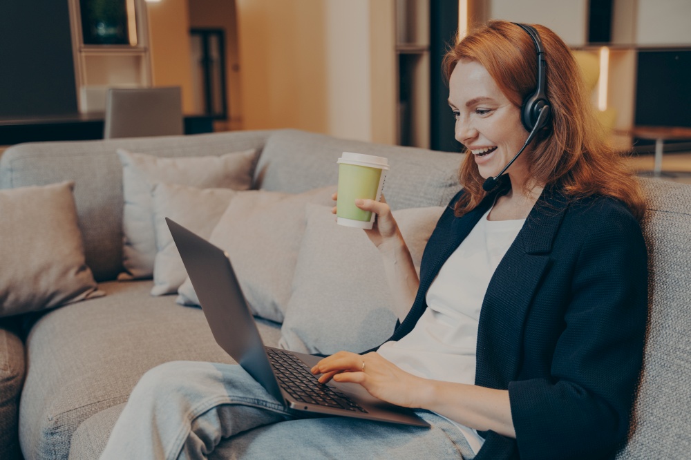 Excited red haired smiling woman with wireless headset and laptop in front of her having online call with colleagues while sitting on sofa in cafe in relaxed atmosphere, holding cup of coffee in hand. Excited red haired smiling woman with wireless headset and laptop sitting on sofa in cafe