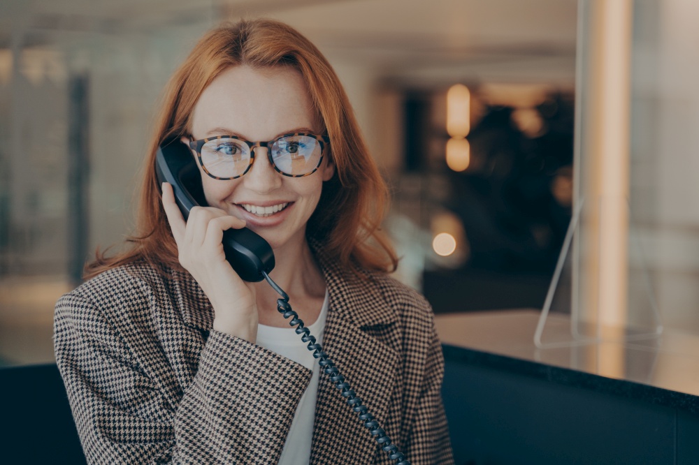 Smiling female office worker with red hair in plaid jacket wearing glasses, talking on landline phone with her co-worker about project completion date, office environment in blurred background. Female office worker in spectacles talking on landline phone with co-worker and smiling at camera