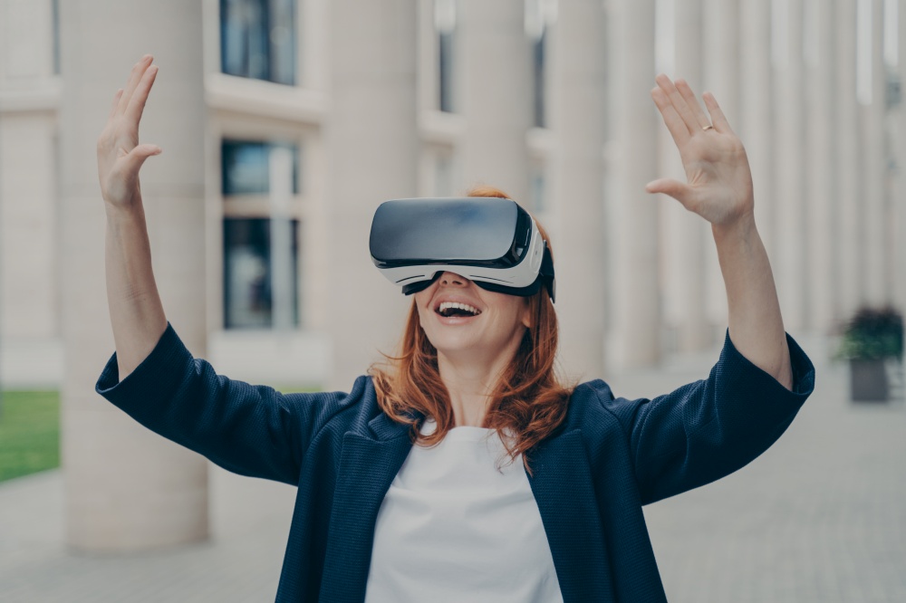 New age of business. Impressed ginger business woman testing virtual reality, using mobile VR headset with excited face expression, standing alone on city street in front of office buildings. Impressed ginger business woman testing virtual reality, using mobile VR headset outside