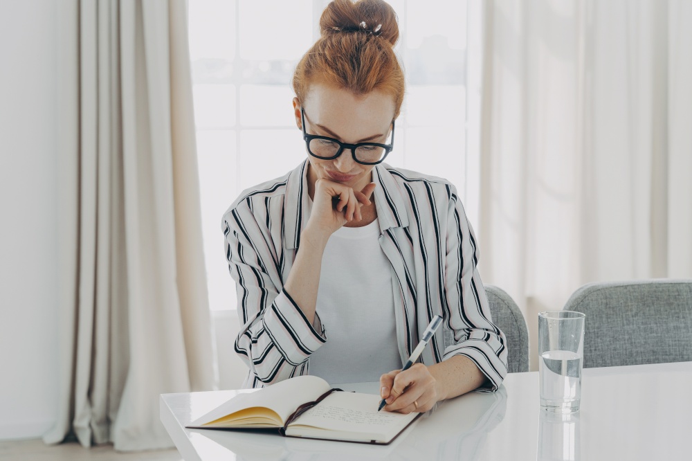 Redhead woman in striped shirt takes notes writes down information in notebook counts month spendings focused in notebook sits at table drinks fresh water cozy domestic interior plans schedule. Redhead woman in striped shirt takes notes writes down information in notebook