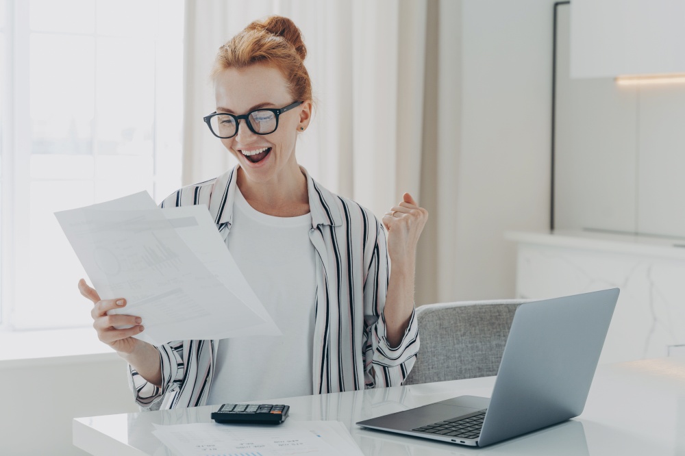 Emotional happy young ginger woman in eyewear holding paper with last mortgage payment banking notification, raising hand with clenched fist, reading document about tax refund from bank. Emotional happy young ginger woman holding paper with last mortgage payment banking notification