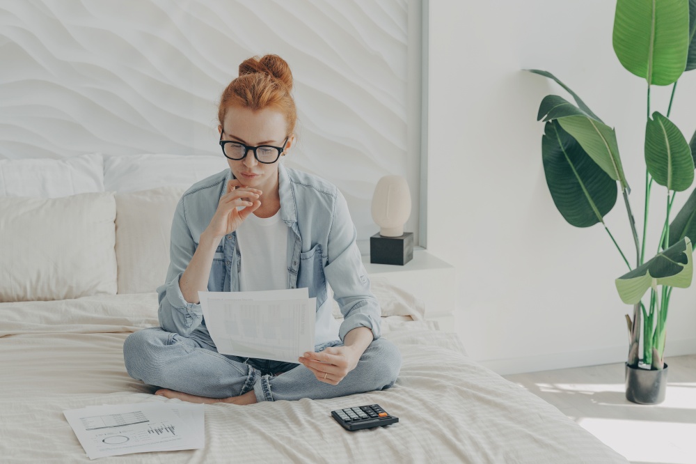 Focused ginger european woman sitting on bed and calculating domestic bills at home, holding papers, reading bank notification while accounting monthly budget and planning family expenses. Focused ginger european woman sitting on bed and calculating domestic bills at home, holding papers
