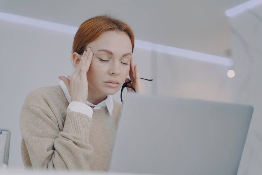 Sick young woman has temples pain in office. Depressed employee has headache. Exhausted girl is holding her head with hands. Concept of overworking and pressure at workplace.. Sick young woman has temples pain in office. Employee has headache at workplace.