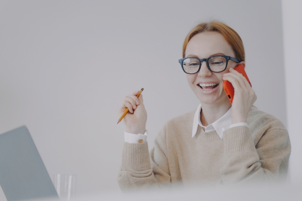 Young business woman is talking on telephone in office. Happy girl talking on cell phone to friend and laughing. Phone conference. Information and communication concept.. Young business woman is talking on telephone and laughing. Phone conference and communication.