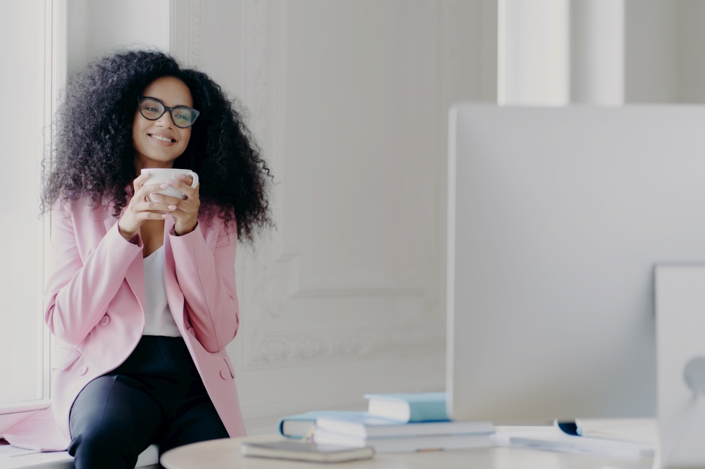 Joyful curly haired female freelancer or blogger has coffee break, holds white mug with beverage, poses near window, focused in computer, watches useful training video, wears near formal clothes