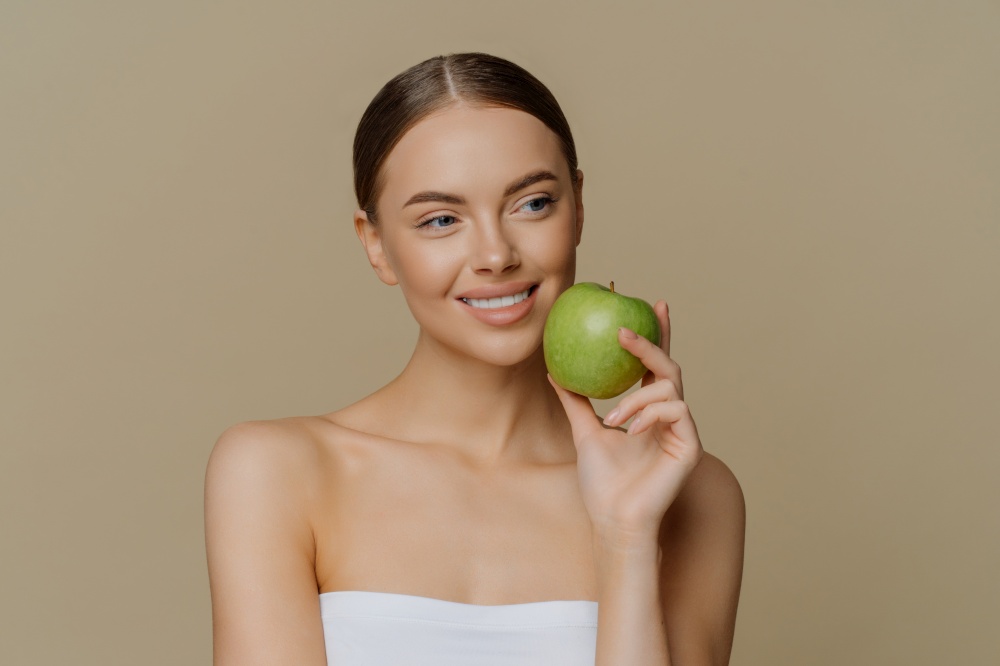 Thoughtful charming European woman holds apple near face smiles gently has white perfect teeth healthy clean skin wrapped in shower towel stands with bare shoulders against brown background.