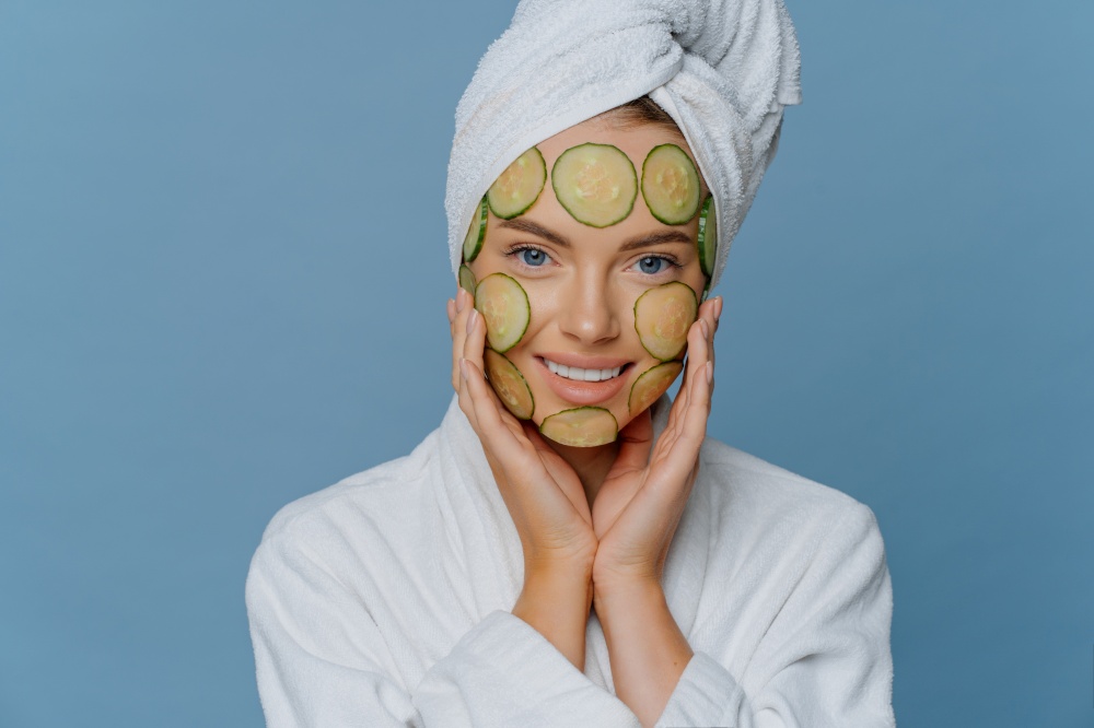 Young beautiful woman applies cucumber slices on face has perfect clean skin touches cheeks gently dressed in comfortable dressing gown has towel wrapped on head. Cosmetology and spa concept