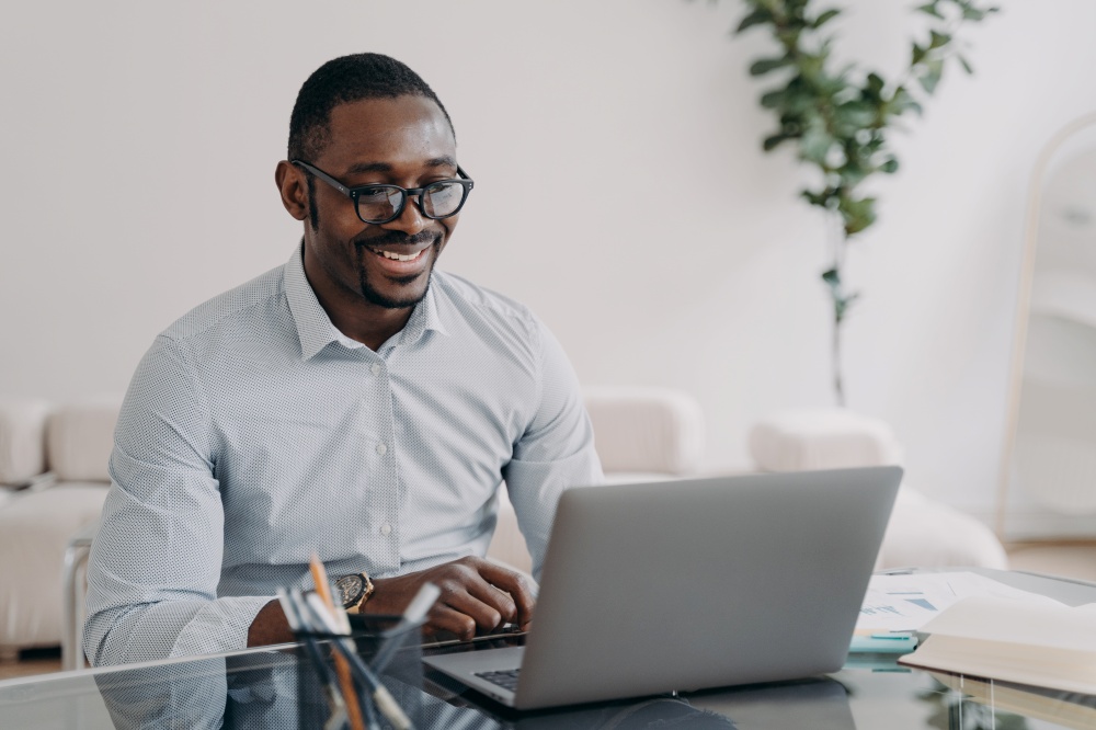 Smiling african american businessman wearing glasses working at laptop, makes successful deal. Happy black man employee glad to receive good news looking at computer screen at office desk.. Smiling african american man wearing glasses working on business project at laptop at office desk